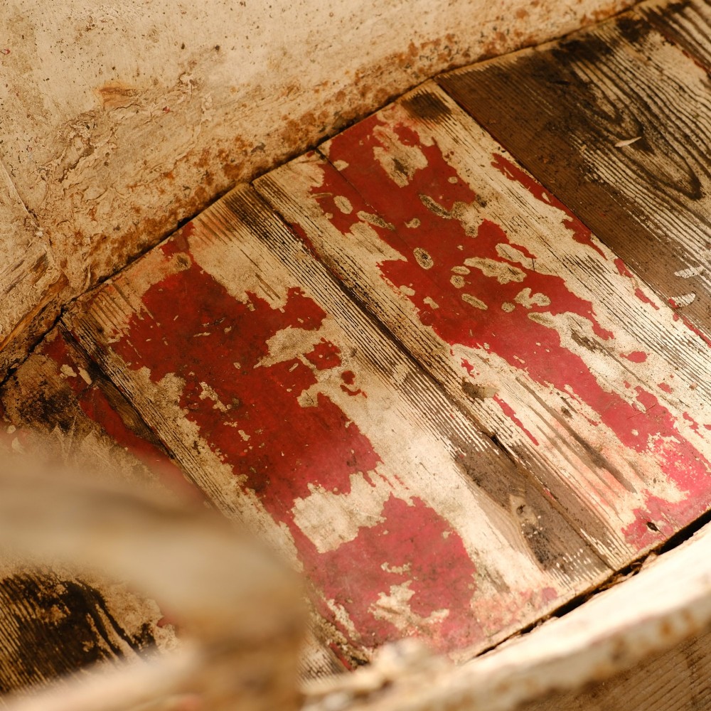 A weathered painted metal fairground swing boat featuring a red wooden seat, riveted sheet body, and curved iron safety rails. View XIII
