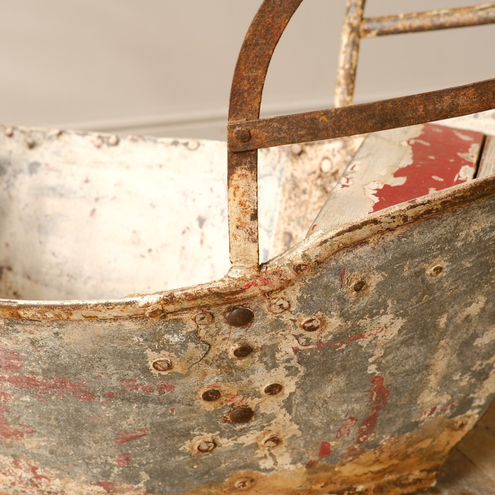 A weathered painted metal fairground swing boat featuring a red wooden seat, riveted sheet body, and curved iron safety rails. View XIV