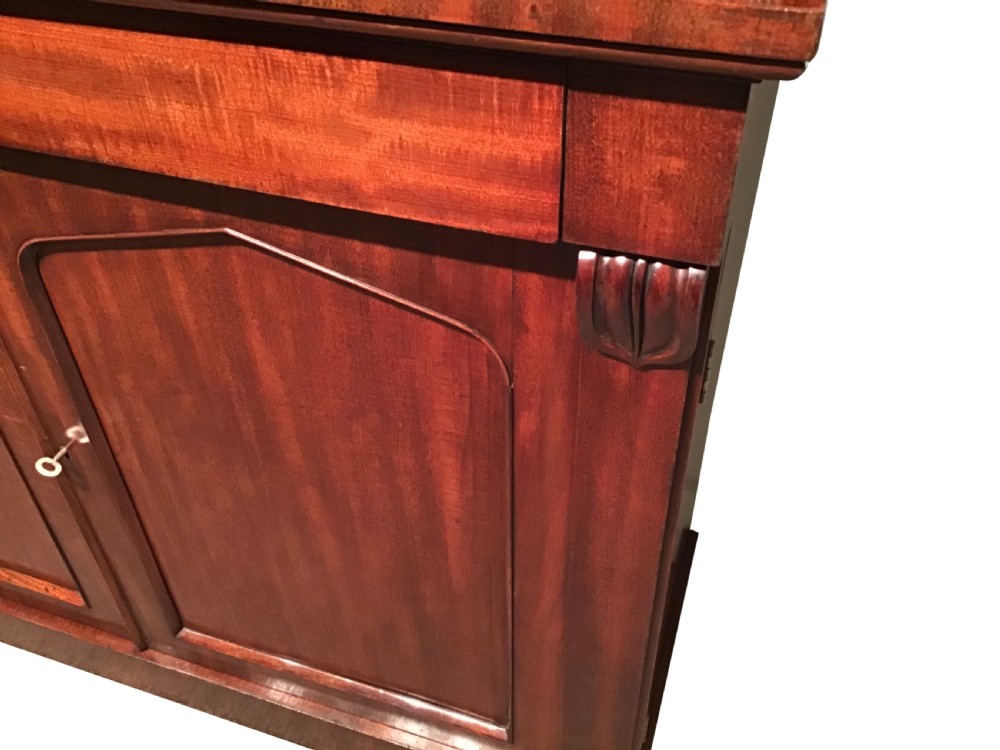 An antique flame mahogany side cabinet featuring an upper shelf supported by scrolled brackets above two panelled cupboard doors. View II