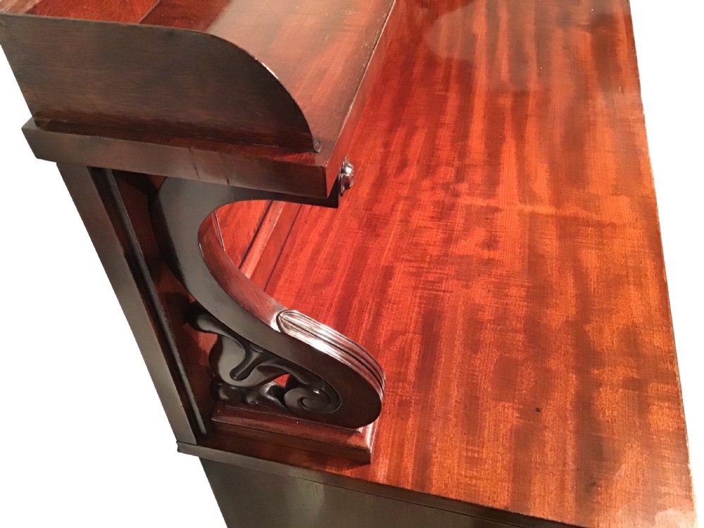An antique flame mahogany side cabinet featuring an upper shelf supported by scrolled brackets above two panelled cupboard doors. View III