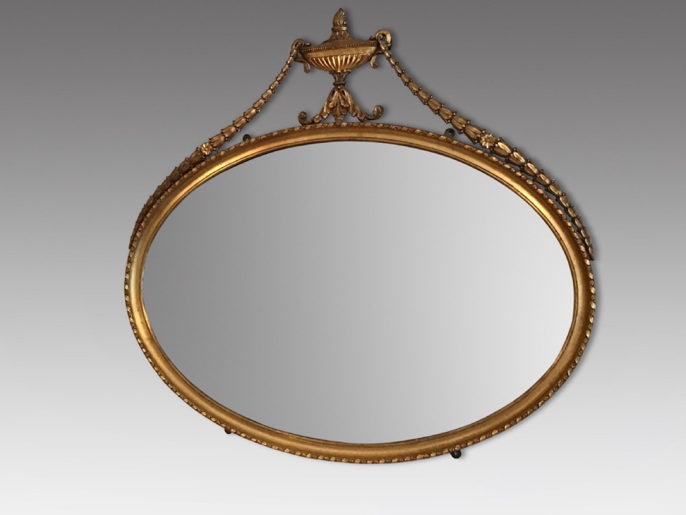 An oval gilded wall mirror featuring an ornamental top urn cresting flanked by draped leaf garlands and a beaded frame border. View II