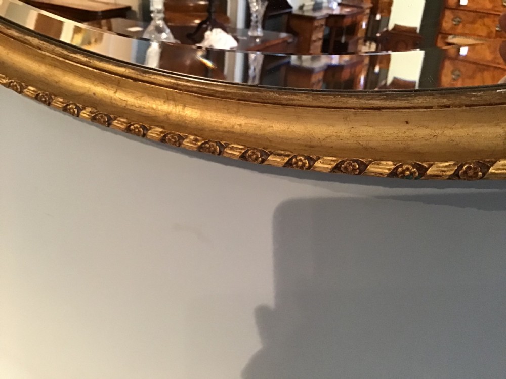 An oval gilded wall mirror featuring an ornamental top urn cresting flanked by draped leaf garlands and a beaded frame border. View III