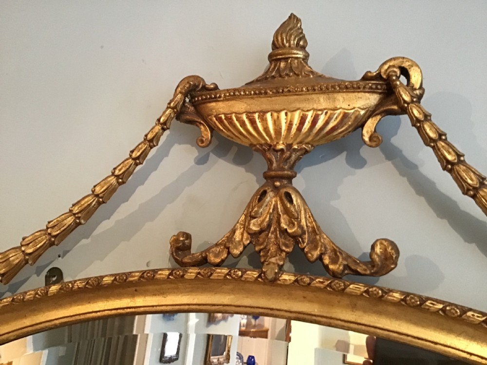 An oval gilded wall mirror featuring an ornamental top urn cresting flanked by draped leaf garlands and a beaded frame border. View IV