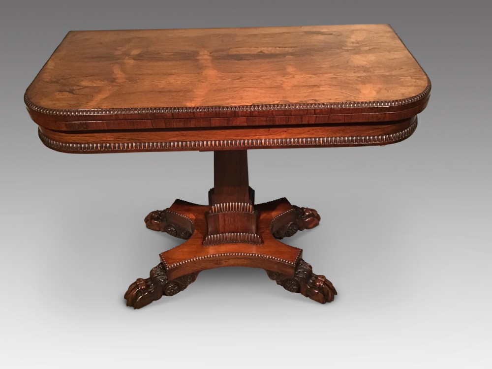 A William IV rosewood fold-over card table featuring a rectangular top with rounded corners, gadrooned edging, a central pedestal stem, and a carved quadform base resting on paw feet.