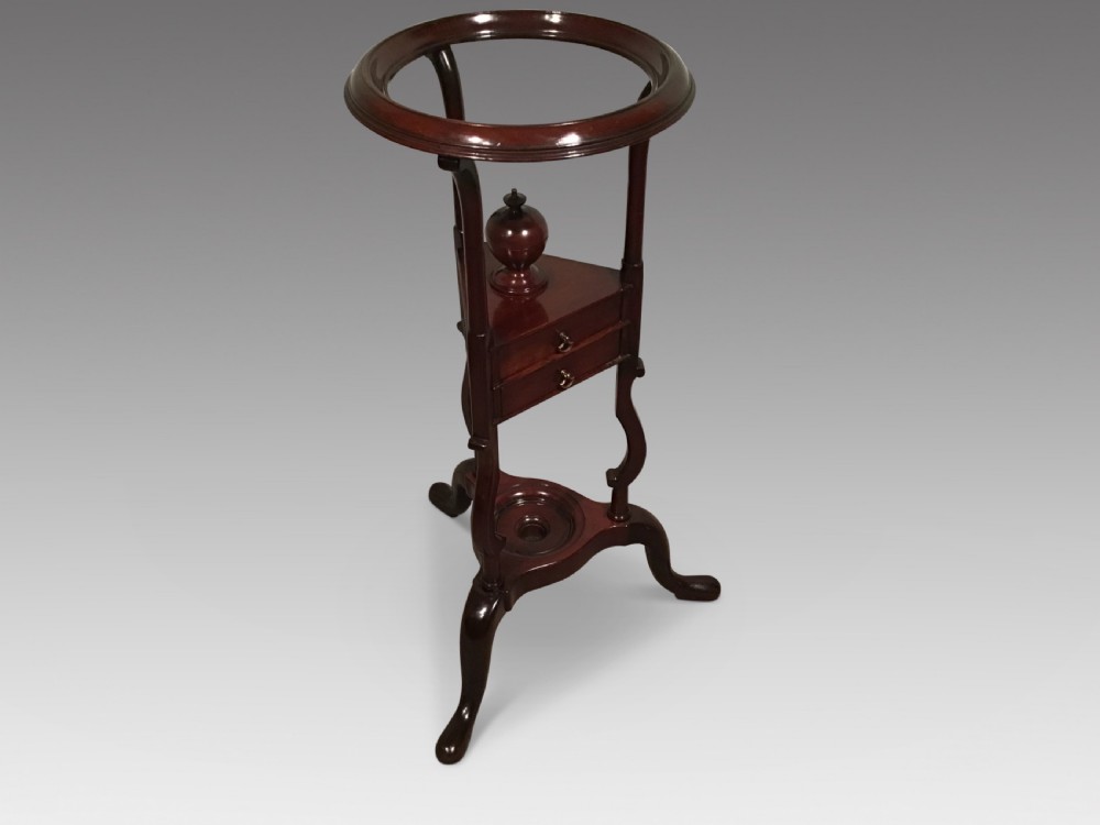 A dark mahogany Georgian wig stand featuring a circular top ring, a triangular drawer section with an urn finial, and a tripod base with cabriole legs.