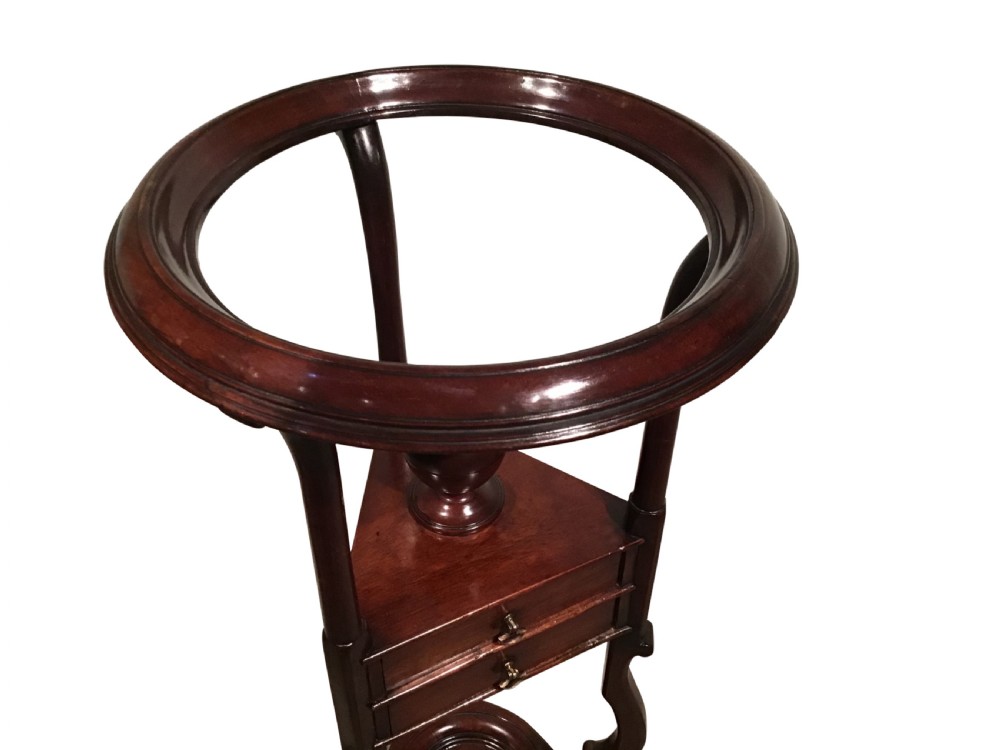 A dark mahogany Georgian wig stand featuring a circular top ring, a triangular drawer section with an urn finial, and a tripod base with cabriole legs. View IV
