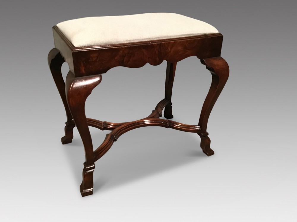 An antique walnut footstool featuring a rectangular cream fabric upholstered seat supported by four cabriole legs and an X-shaped cross stretcher.