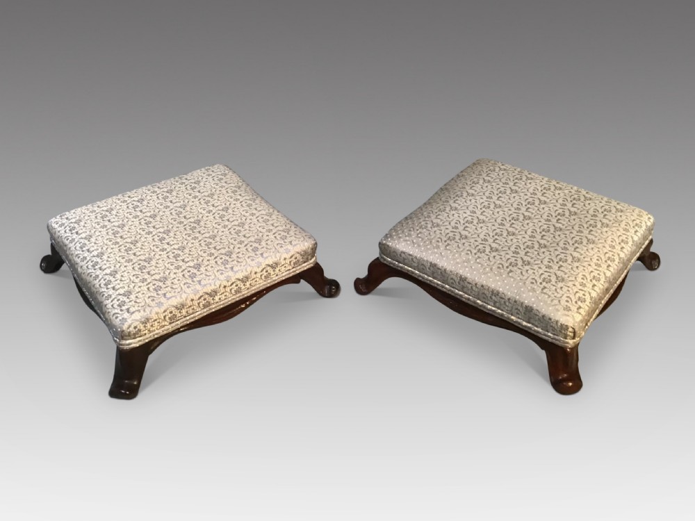 A pair of low antique footstools featuring dark carved mahogany cabriole legs and silver floral patterned upholstery.