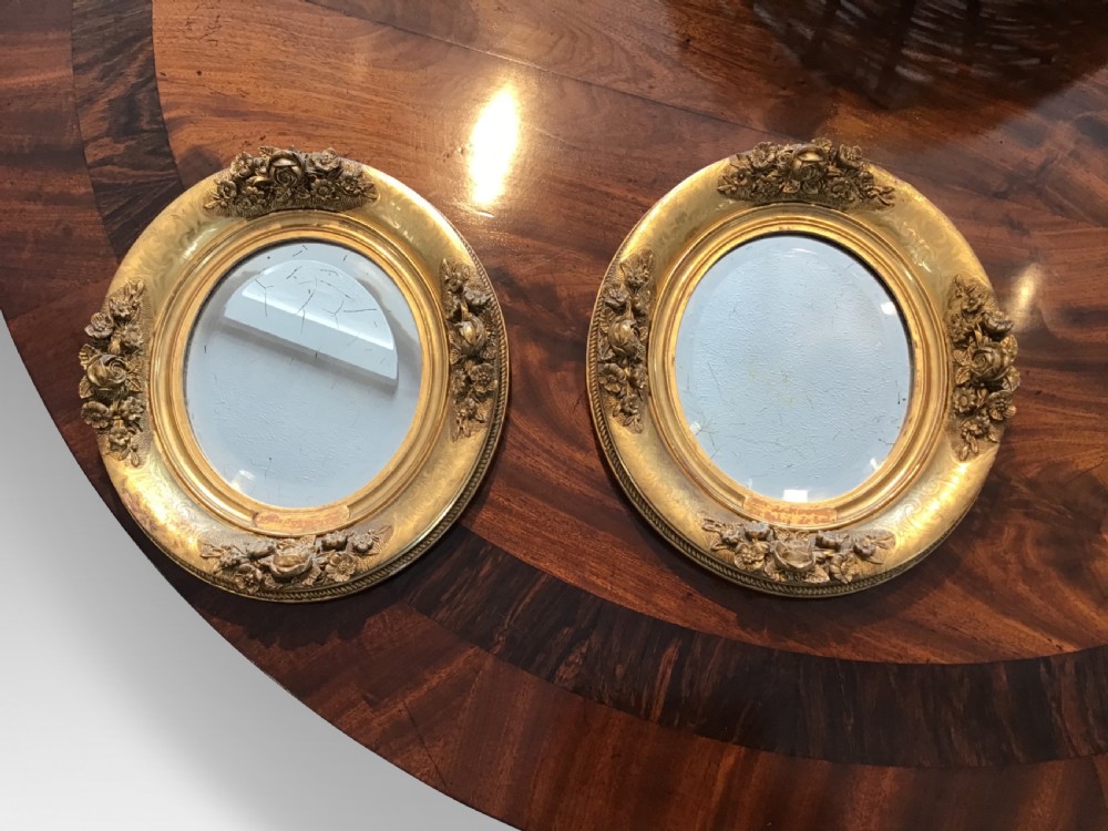 A pair of oval giltwood mirrors featuring detailed floral moulding and engraved detailing along the gilded frames.