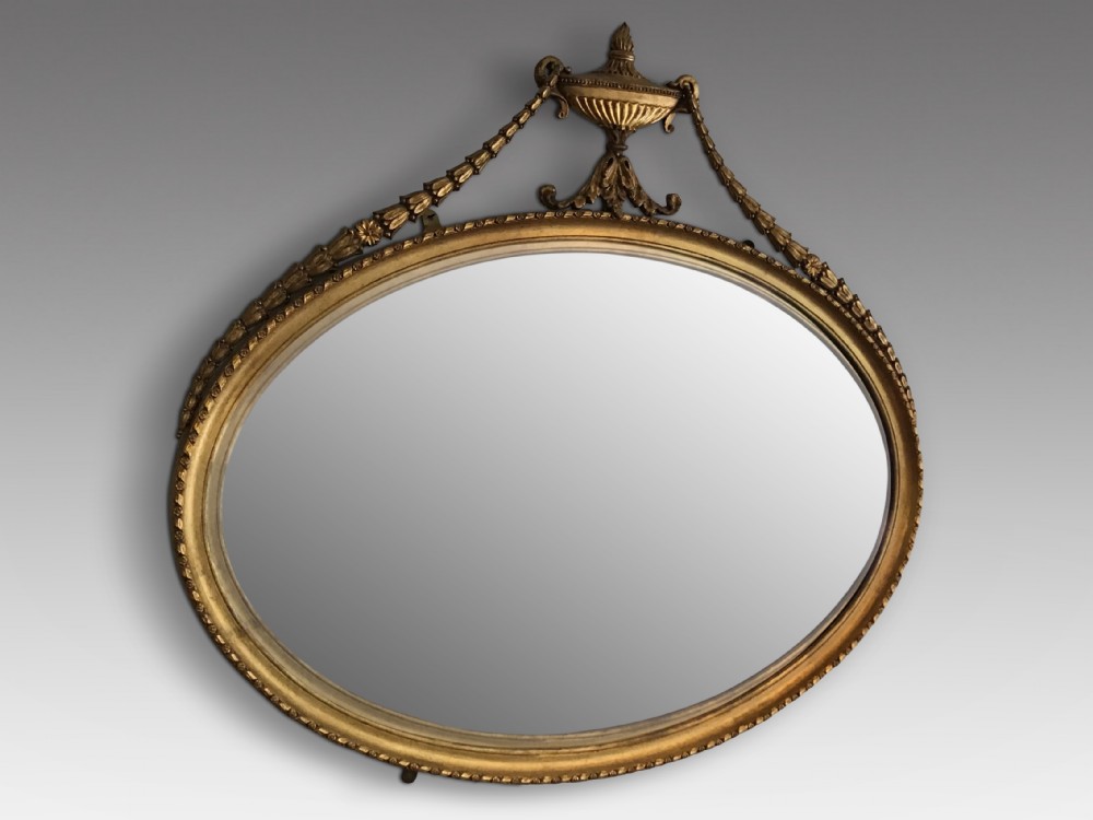 An oval gilded wall mirror featuring an ornamental top urn cresting flanked by draped leaf garlands and a beaded frame border.