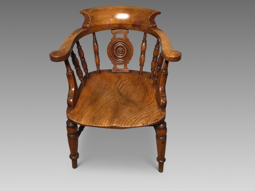 An antique wooden smokers bow armchair with turned spindles, a carved central splat, and a shaped saddle seat.