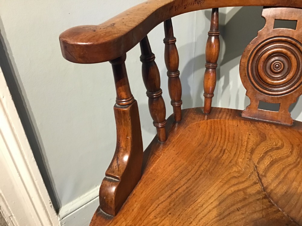 An antique wooden smokers bow armchair with turned spindles, a carved central splat, and a shaped saddle seat. View IV