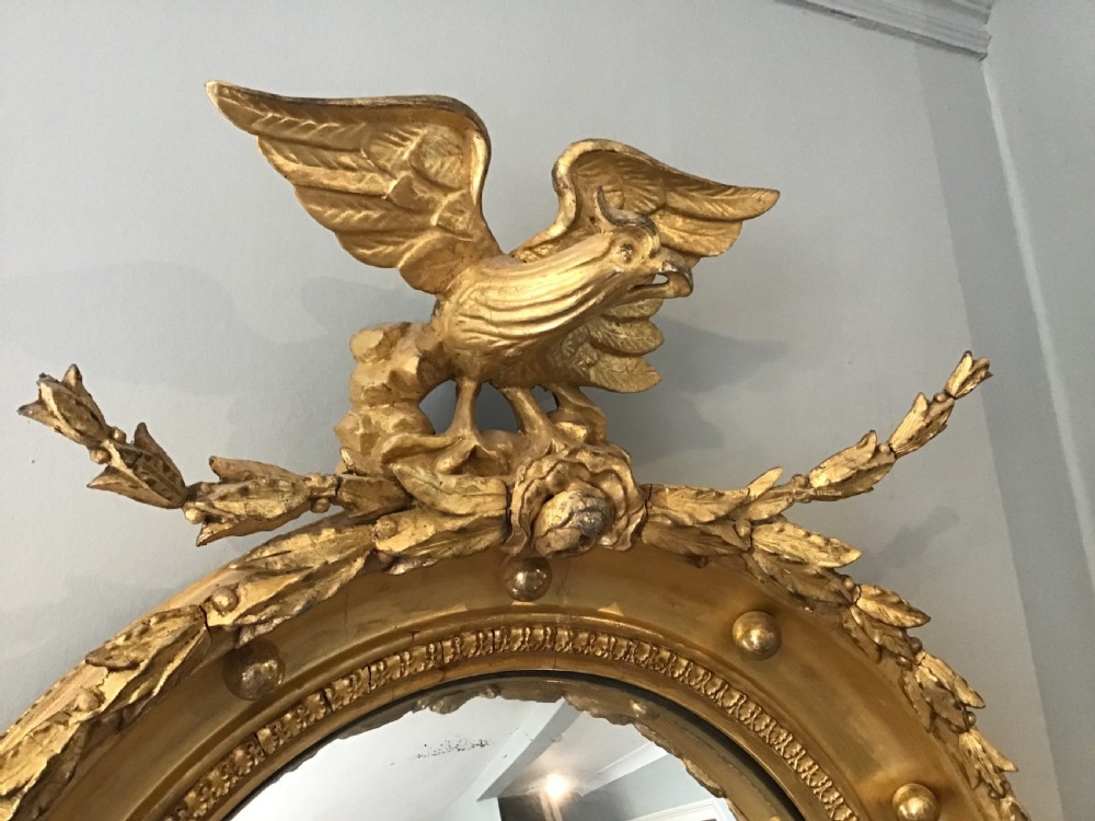 A circular Regency giltwood convex mirror featuring a carved eagle cresting at the top, decorative spheres around the frame rim, and an acanthus leaf pendant below. View II
