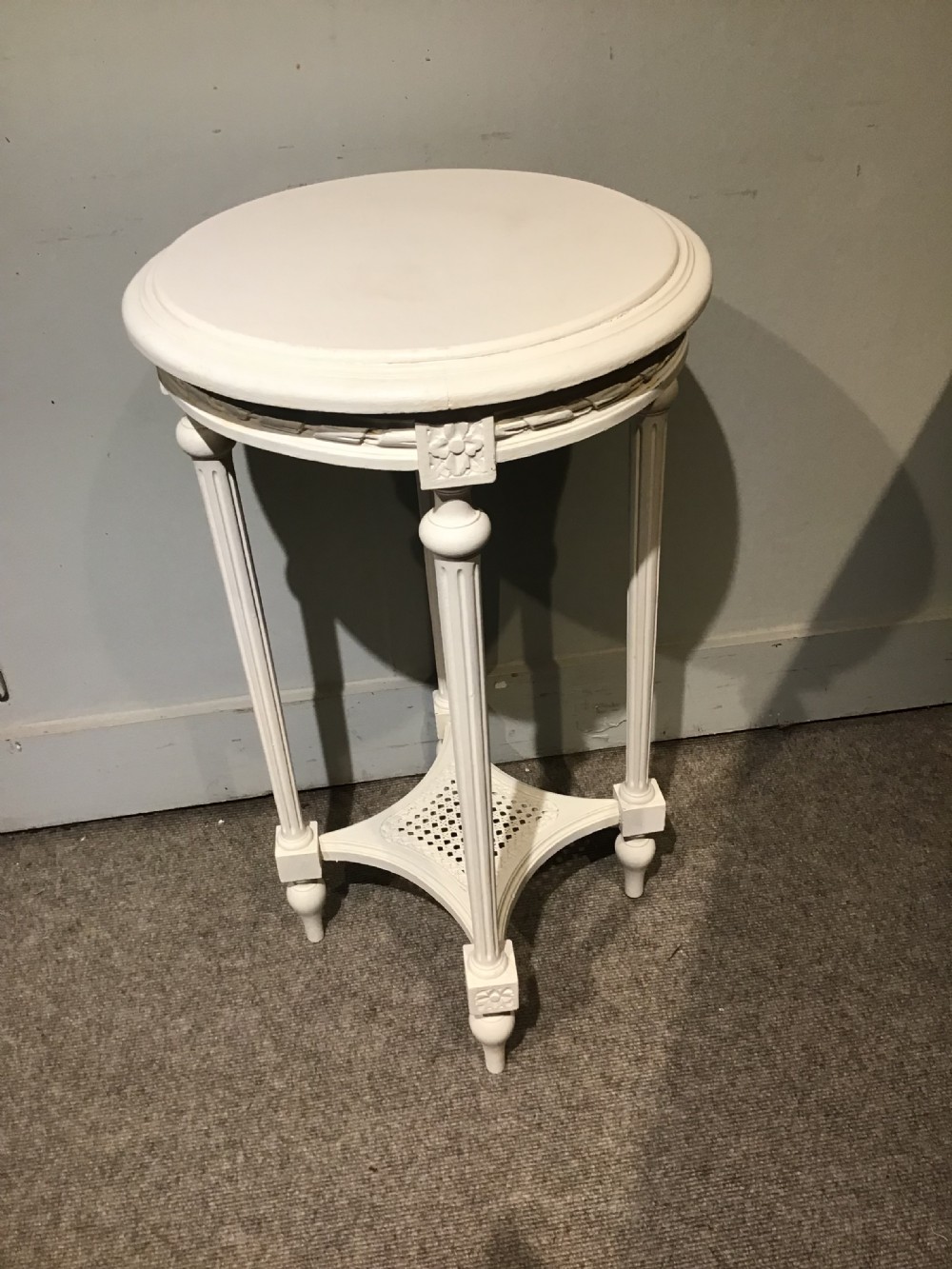 A circular off-white painted side table featuring four fluted legs, carved rosette blocks, and a lower square cane shelf.