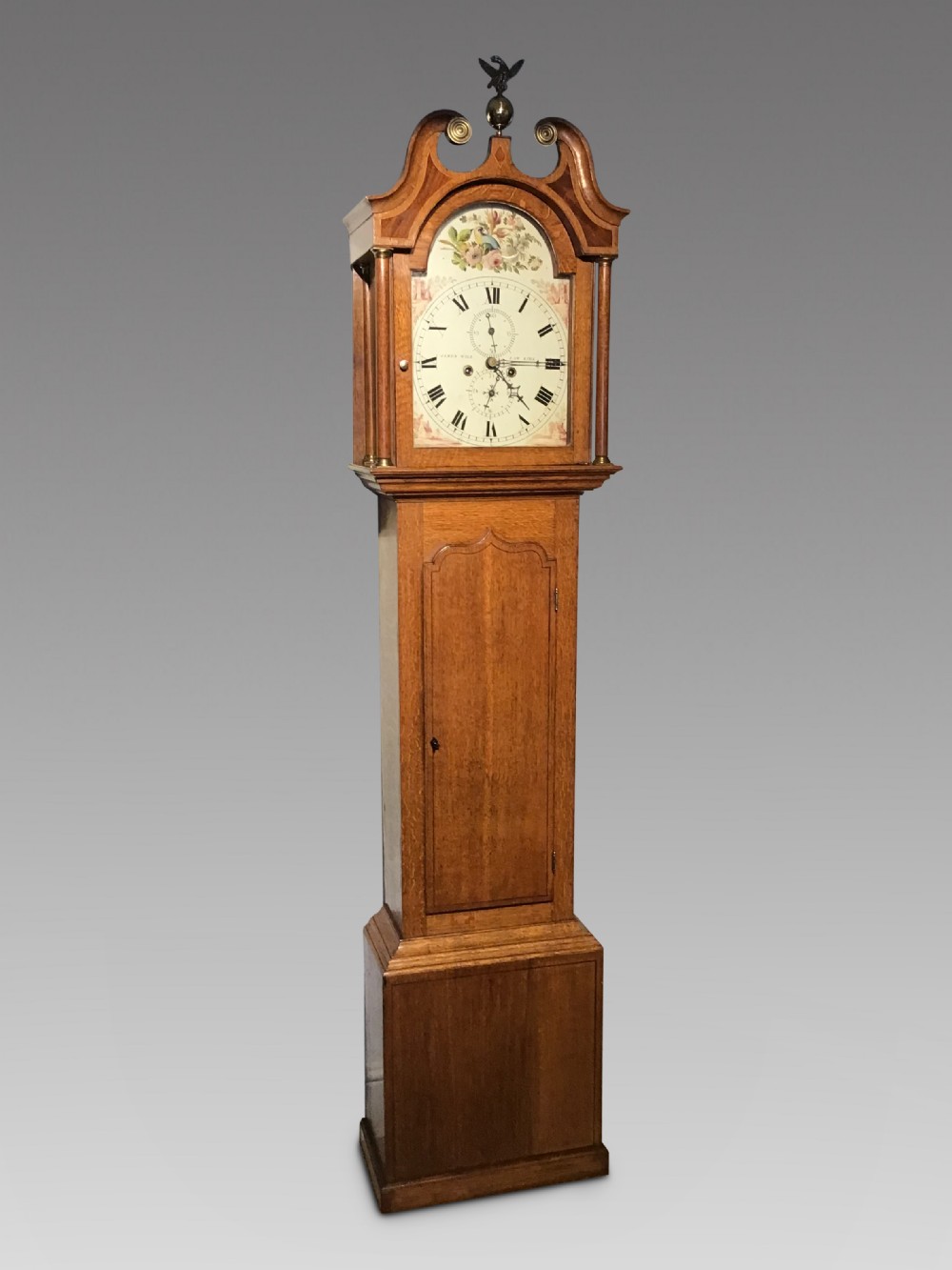 An antique oak longcase clock featuring a painted dial with Roman numerals, a swan-neck pediment with a brass finial, and a wooden trunk with an arched door.