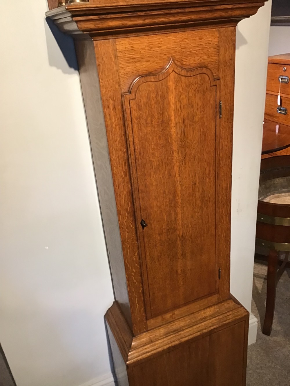 An antique oak longcase clock featuring a painted dial with Roman numerals, a swan-neck pediment with a brass finial, and a wooden trunk with an arched door. View III