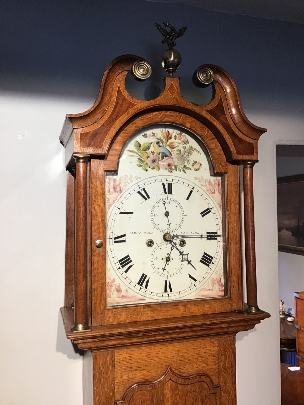 An antique oak longcase clock featuring a painted dial with Roman numerals, a swan-neck pediment with a brass finial, and a wooden trunk with an arched door. View V