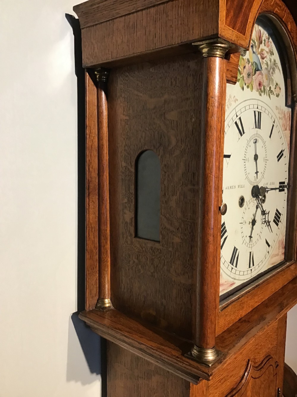 An antique oak longcase clock featuring a painted dial with Roman numerals, a swan-neck pediment with a brass finial, and a wooden trunk with an arched door. View VII