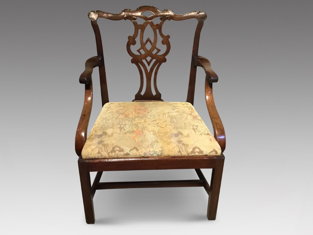 An antique mahogany elbow chair featuring a carved pierced back splat, curved arms, and a soft beige floral tapestry seat.