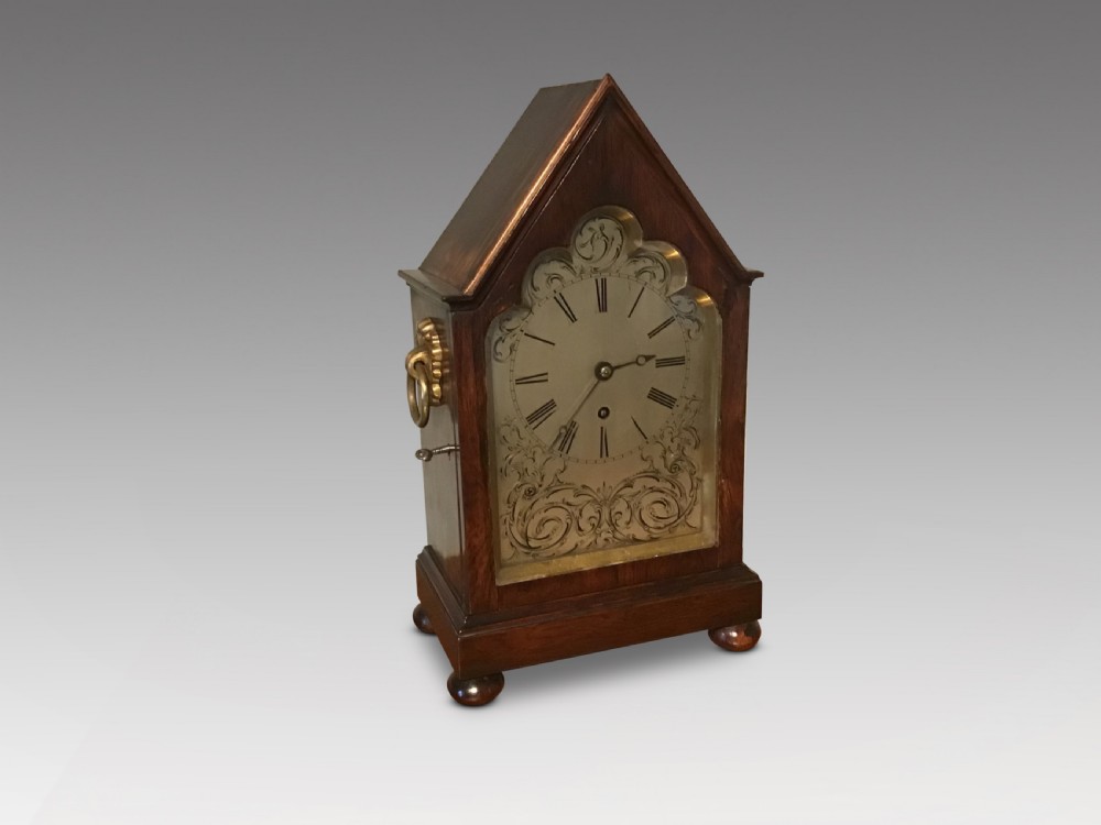 An antique mahogany bracket clock featuring a pointed gabled top, side brass ring handles, ball feet, and an engraved metal dial with Roman numerals.