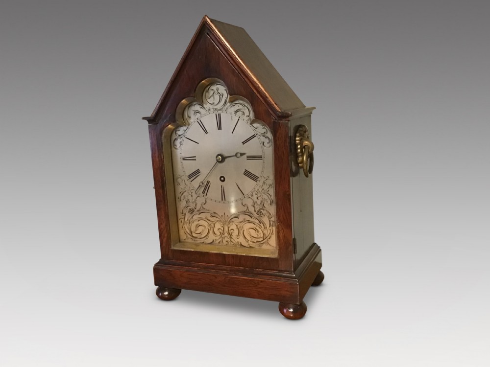 An antique mahogany bracket clock featuring a pointed gabled top, side brass ring handles, ball feet, and an engraved metal dial with Roman numerals. View II