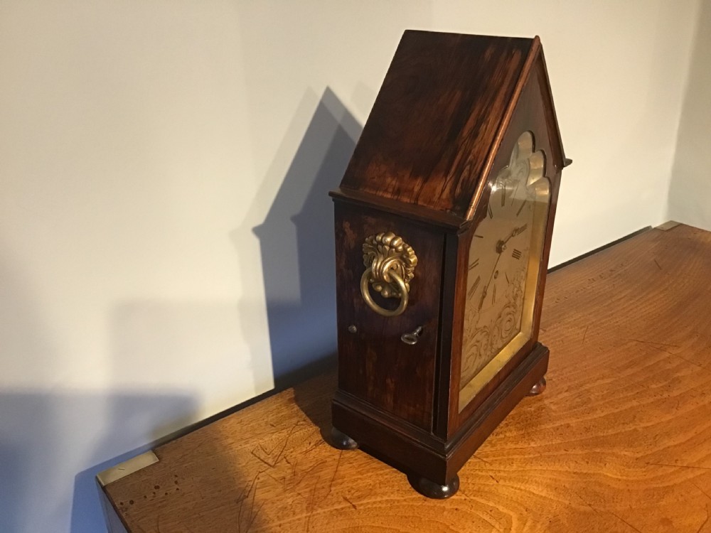An antique mahogany bracket clock featuring a pointed gabled top, side brass ring handles, ball feet, and an engraved metal dial with Roman numerals. View V