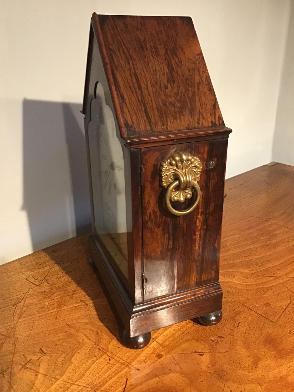 An antique mahogany bracket clock featuring a pointed gabled top, side brass ring handles, ball feet, and an engraved metal dial with Roman numerals. View VI