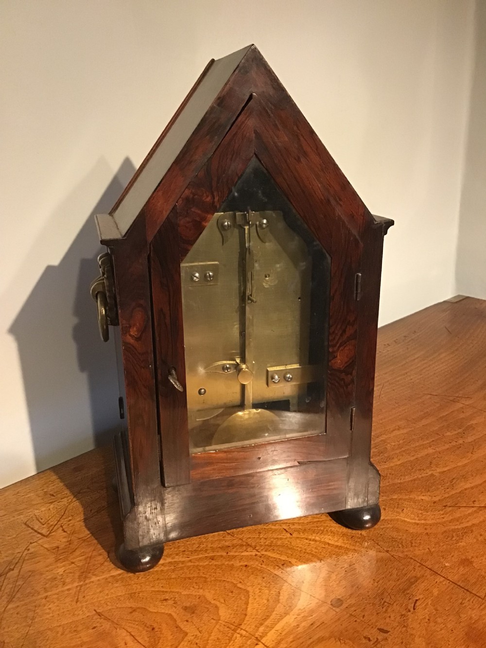 An antique mahogany bracket clock featuring a pointed gabled top, side brass ring handles, ball feet, and an engraved metal dial with Roman numerals. View VII