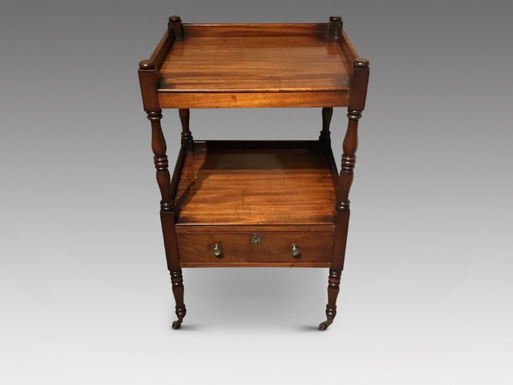 A warm brown mahogany tiered side table with turned supports, a gallery top, an open shelf, a lower drawer with brass knobs, and brass castors.
