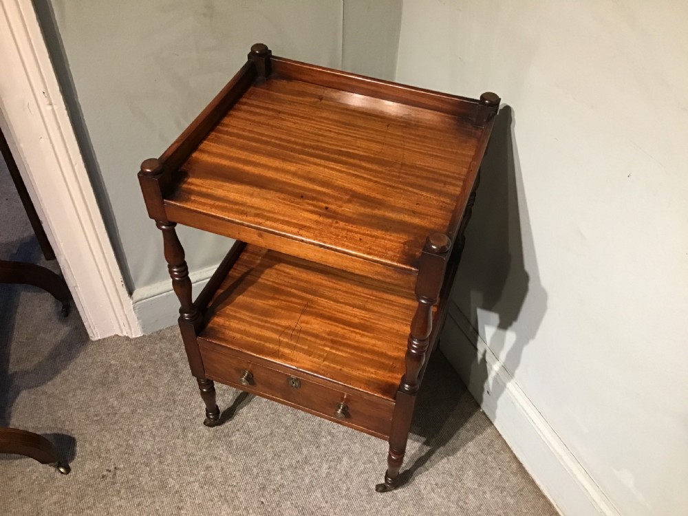 A warm brown mahogany tiered side table with turned supports, a gallery top, an open shelf, a lower drawer with brass knobs, and brass castors. View II