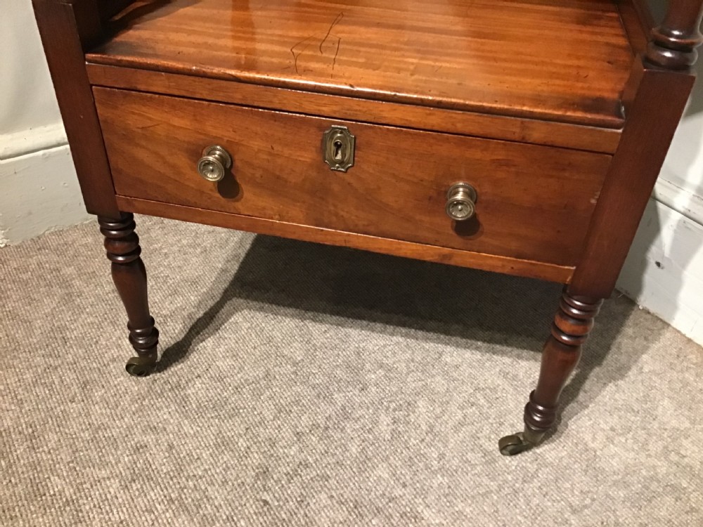 A warm brown mahogany tiered side table with turned supports, a gallery top, an open shelf, a lower drawer with brass knobs, and brass castors. View III
