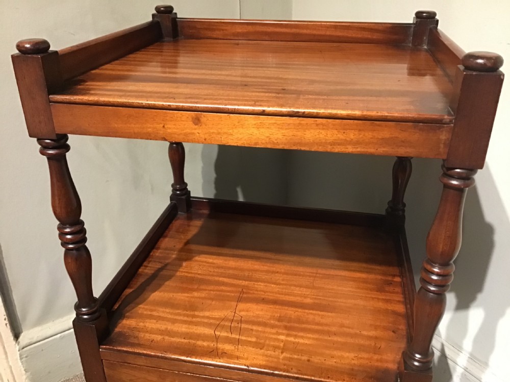 A warm brown mahogany tiered side table with turned supports, a gallery top, an open shelf, a lower drawer with brass knobs, and brass castors. View IV