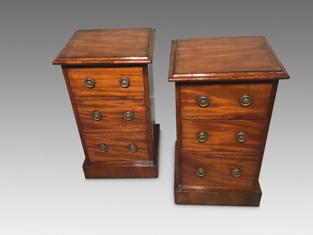 A pair of narrow antique mahogany bedside chests, each featuring three drawers with round brass ring handles and a plinth base.