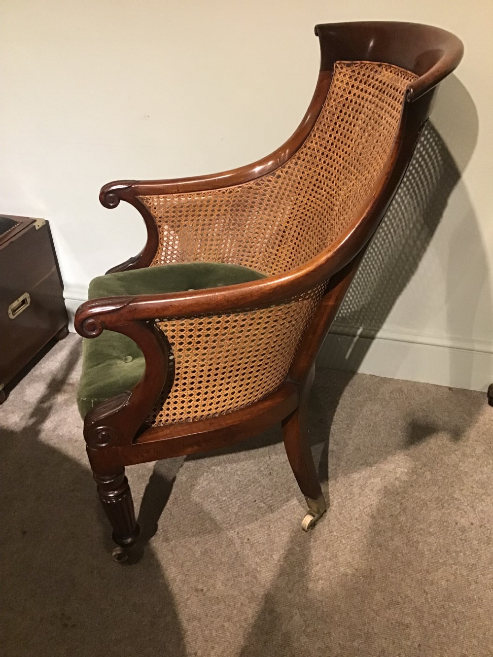 A Regency mahogany bergere armchair featuring hand-woven cane panels, reeded turned front legs on brass castors, and a buttoned green velvet seat cushion. View IV