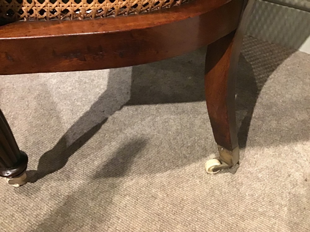 A Regency mahogany bergere armchair featuring hand-woven cane panels, reeded turned front legs on brass castors, and a buttoned green velvet seat cushion. View V