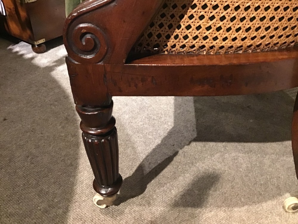 A Regency mahogany bergere armchair featuring hand-woven cane panels, reeded turned front legs on brass castors, and a buttoned green velvet seat cushion. View VI