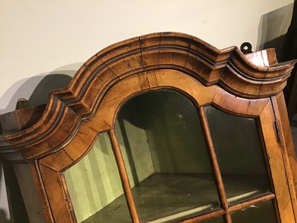 An antique walnut wall-hanging corner cabinet featuring an arched moulded pediment, glazed door with glass panes, and a green-painted interior with shelves. View IV