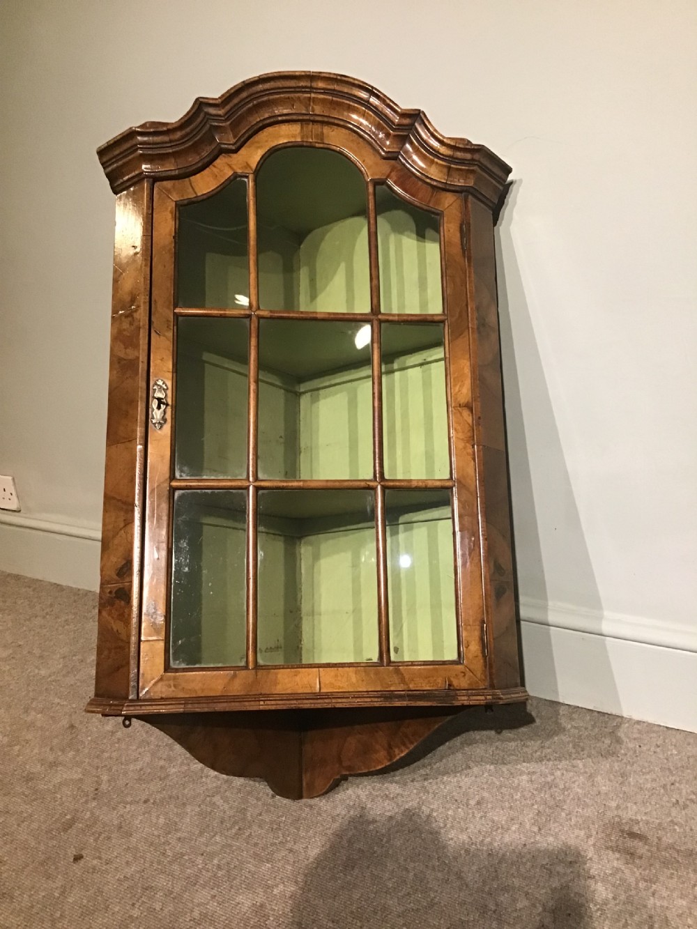 An antique walnut wall-hanging corner cabinet featuring an arched moulded pediment, glazed door with glass panes, and a green-painted interior with shelves. View VII