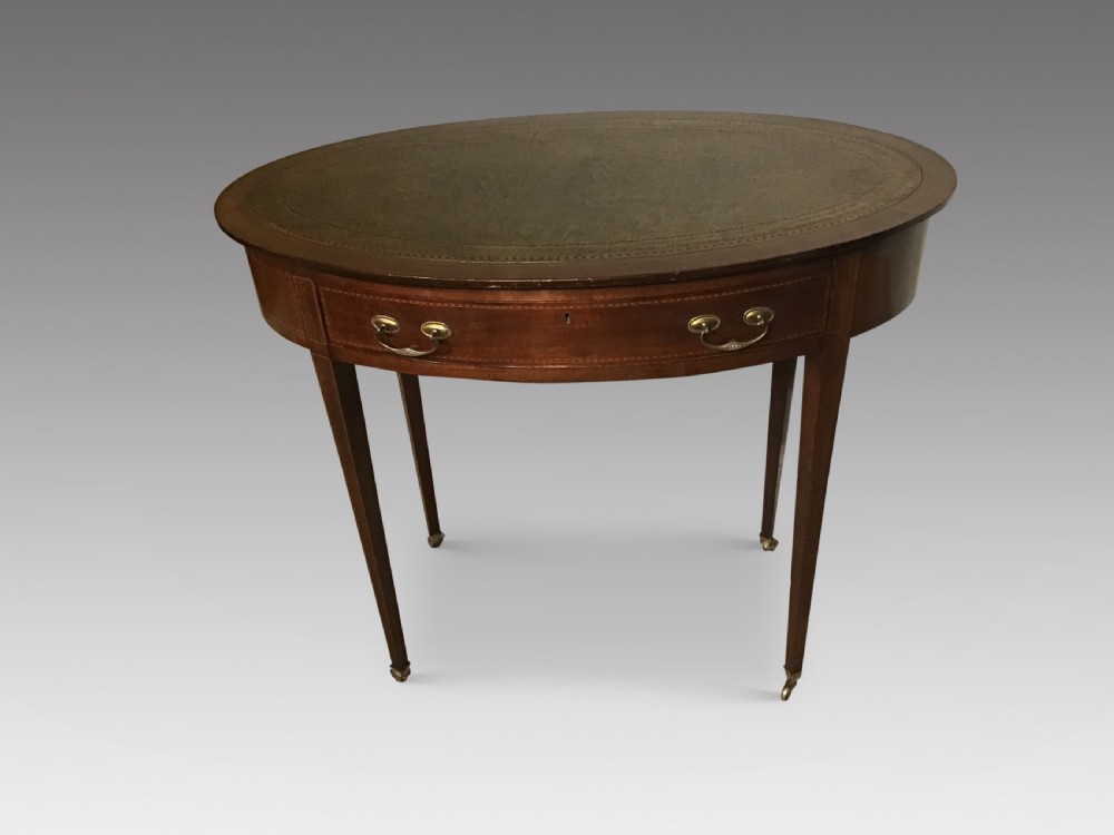 An antique oval mahogany writing table featuring a green leather top with gold tooling, a frieze drawer, and square tapered legs on brass castors.
