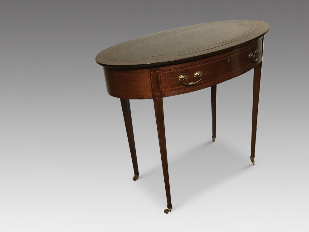 An antique oval mahogany writing table featuring a green leather top with gold tooling, a frieze drawer, and square tapered legs on brass castors. View II