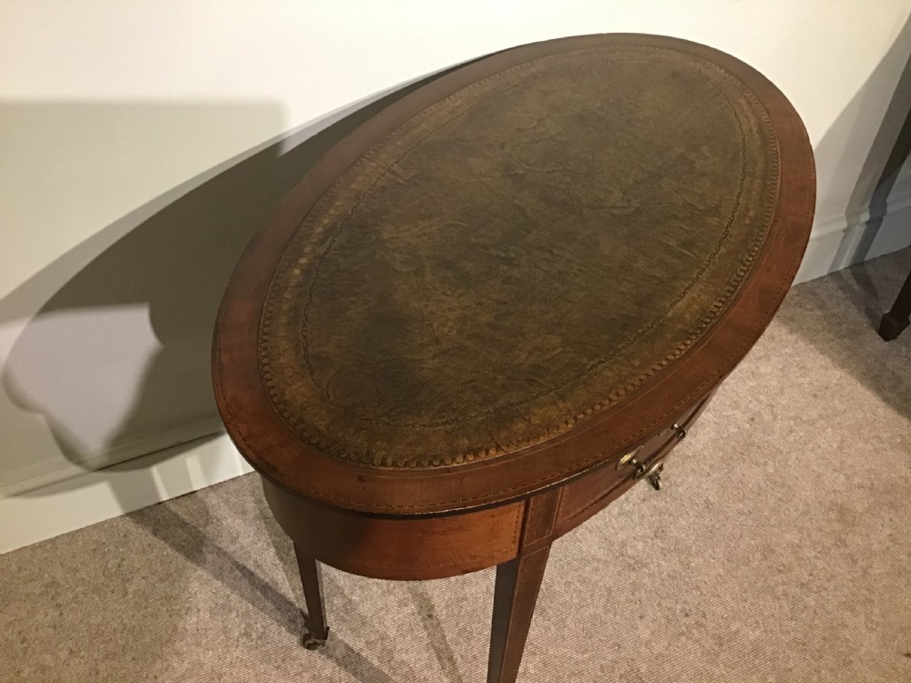An antique oval mahogany writing table featuring a green leather top with gold tooling, a frieze drawer, and square tapered legs on brass castors. View III