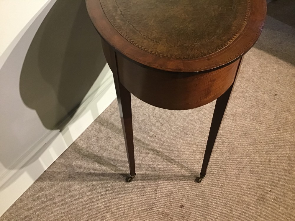 An antique oval mahogany writing table featuring a green leather top with gold tooling, a frieze drawer, and square tapered legs on brass castors. View IV