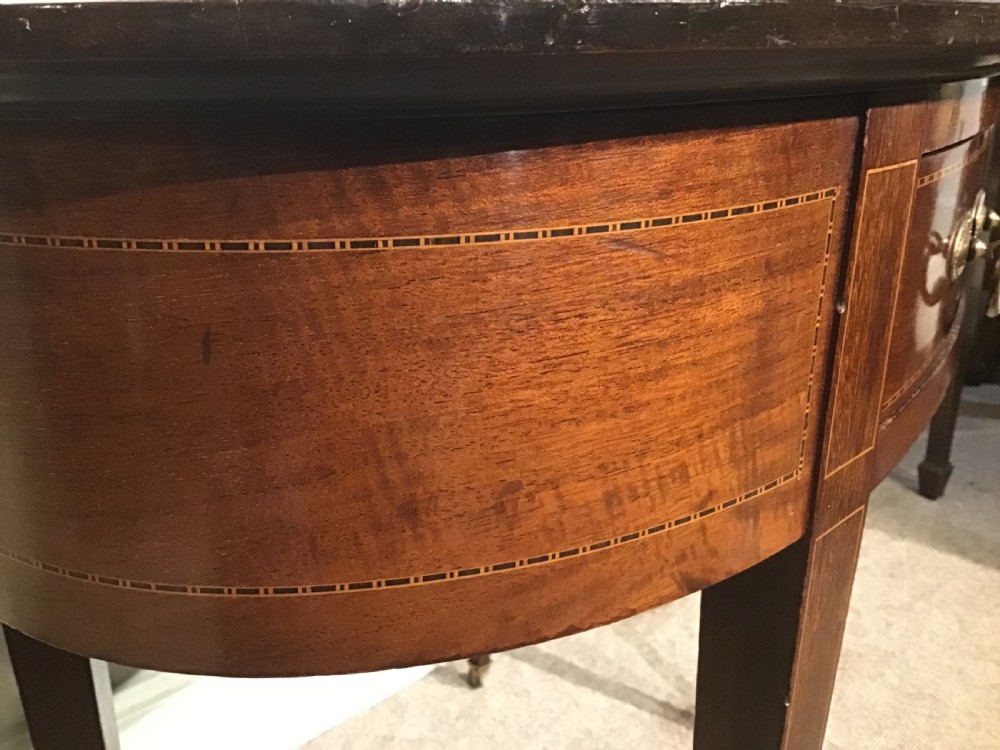 An antique oval mahogany writing table featuring a green leather top with gold tooling, a frieze drawer, and square tapered legs on brass castors. View V