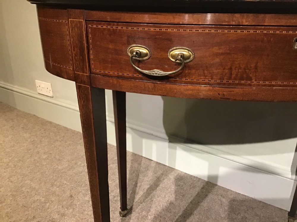 An antique oval mahogany writing table featuring a green leather top with gold tooling, a frieze drawer, and square tapered legs on brass castors. View VI