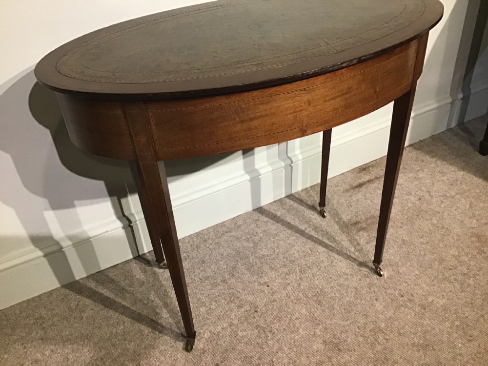 An antique oval mahogany writing table featuring a green leather top with gold tooling, a frieze drawer, and square tapered legs on brass castors. View VIII