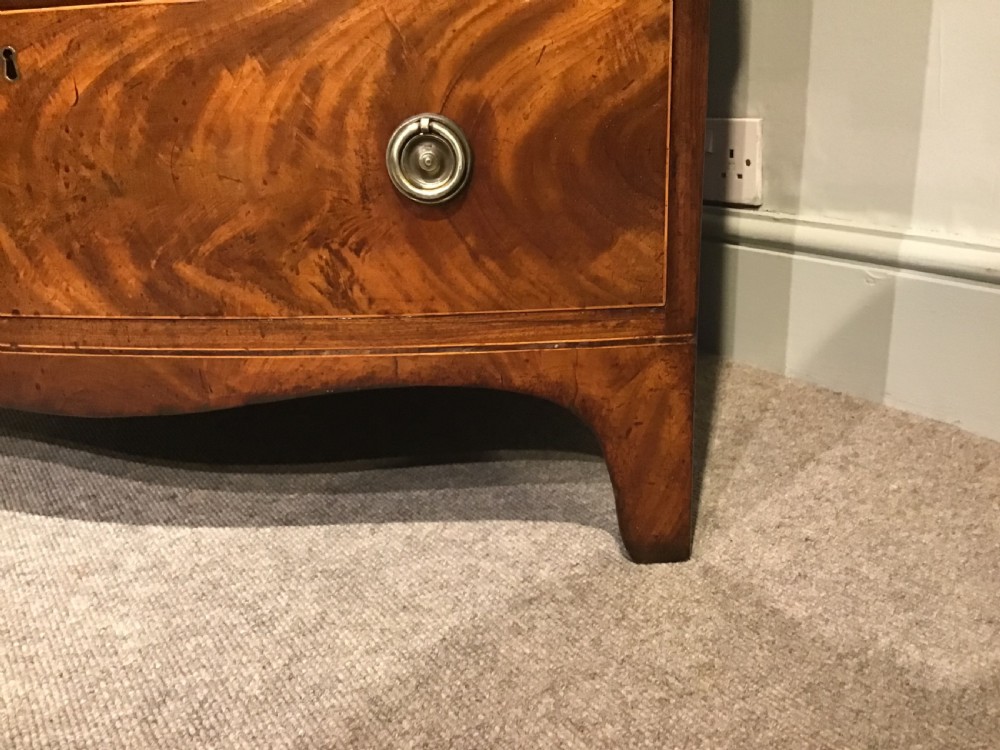A mahogany bow-front chest of drawers featuring four graduated drawers with flame veneers, brass ring handles, and splayed bracket feet. View IV