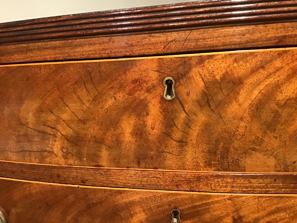 A mahogany bow-front chest of drawers featuring four graduated drawers with flame veneers, brass ring handles, and splayed bracket feet. View V