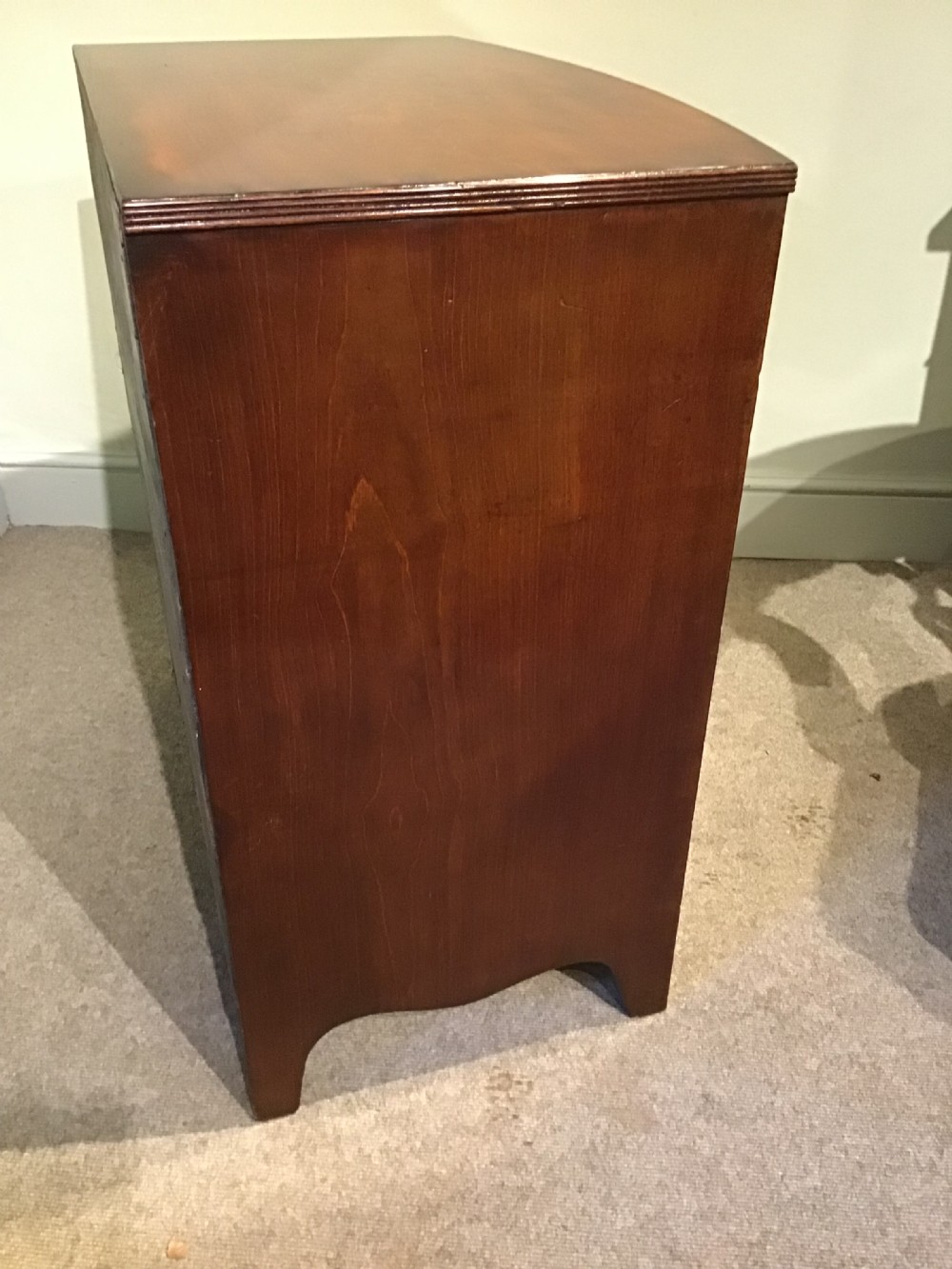 A mahogany bow-front chest of drawers featuring four graduated drawers with flame veneers, brass ring handles, and splayed bracket feet. View X