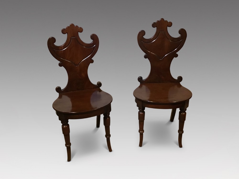A pair of antique dark mahogany hall chairs featuring carved scroll backs, solid wood seats, and turned front legs.