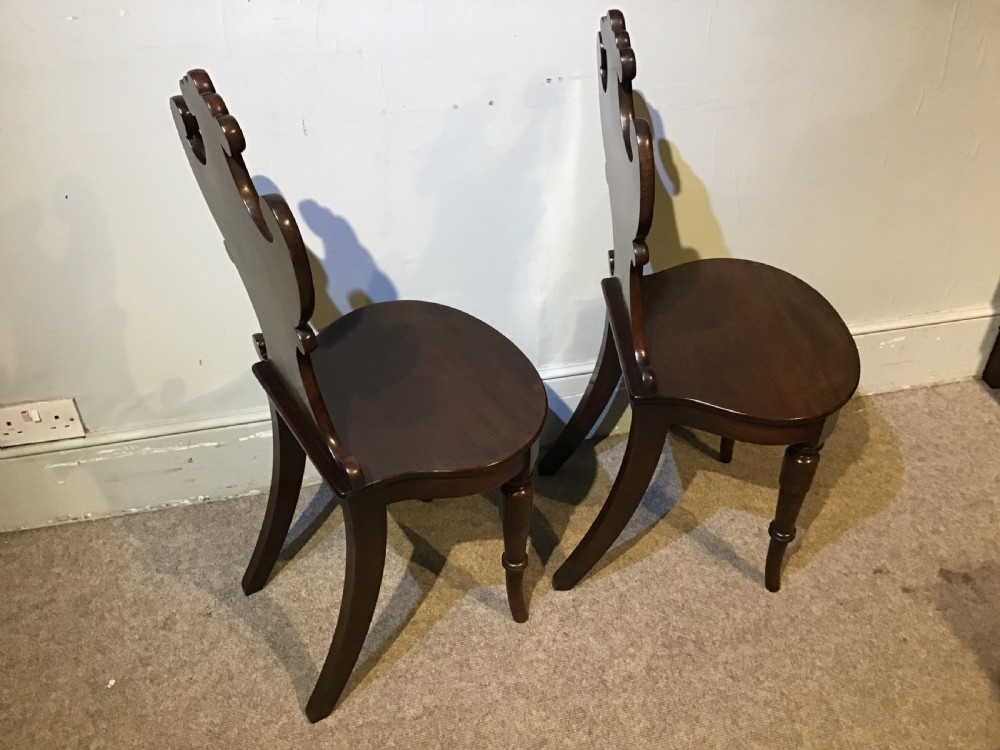 A pair of antique dark mahogany hall chairs featuring carved scroll backs, solid wood seats, and turned front legs. View X